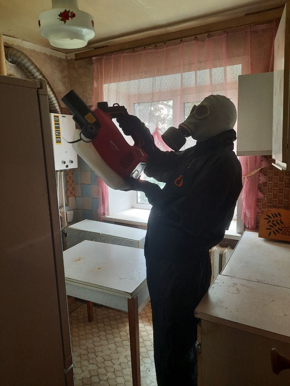 Мастер в защитном костюме и противогазе обрабатывает кухню генератором горячего тумана